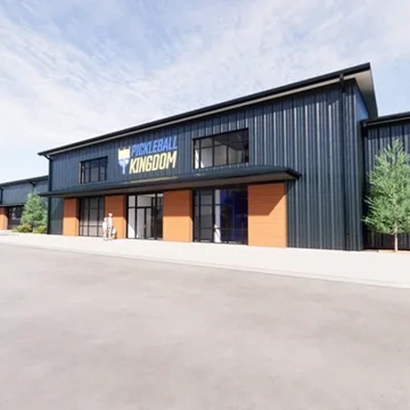 A street-view rendering of the Pickleball Kingdom facility exterior, featuring a modern black metal building with large windows, wood accents, and the Pickleball Kingdom logo displayed above the entrance.