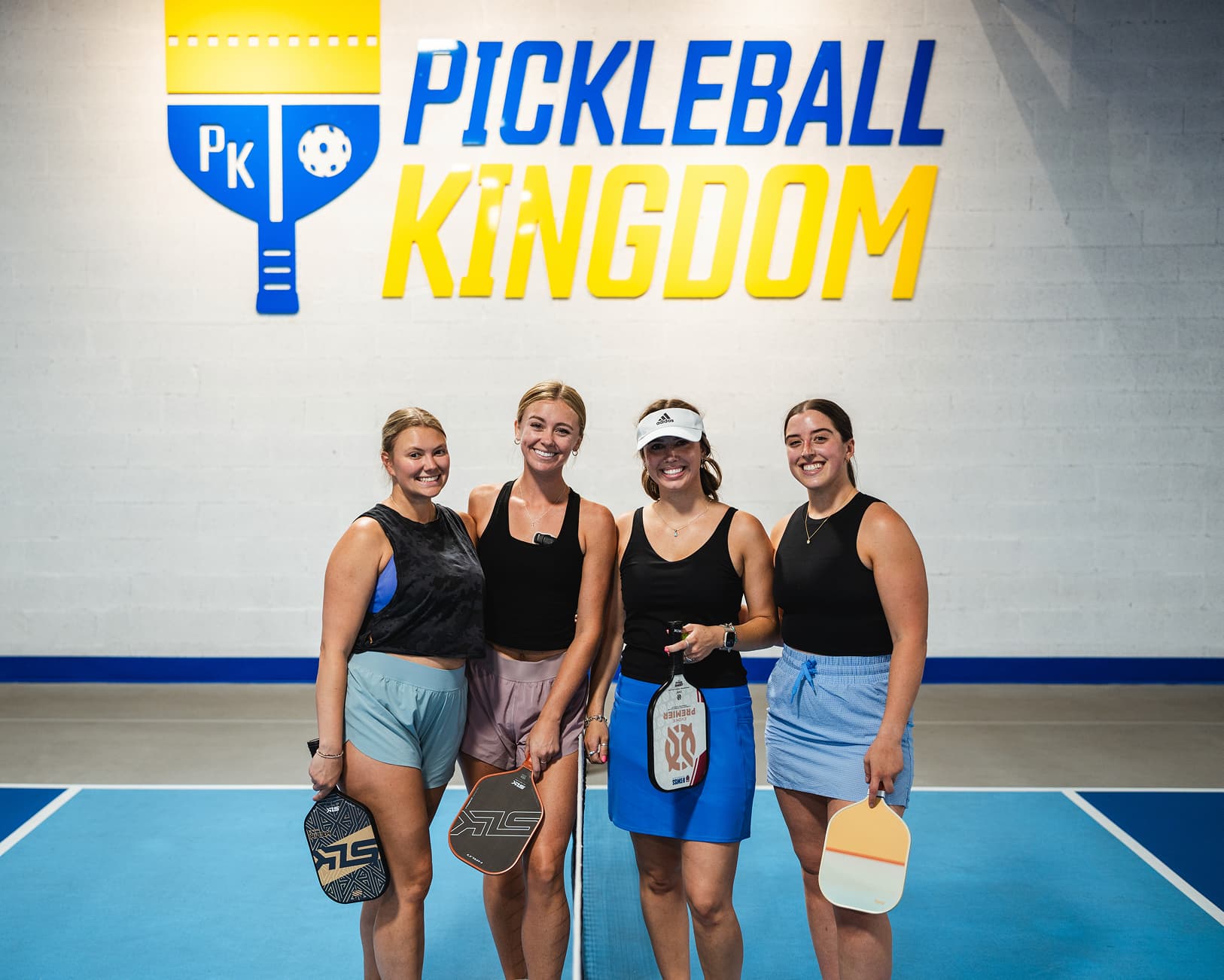 Four women holding paddles and smiling on an indoor pickleball court beneath a large Pickleball Kingdom logo on the wall.