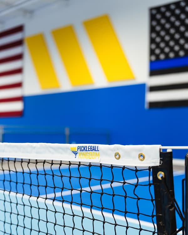 Close-up of a pickleball net with the Pickleball Kingdom logo and bright blue and yellow court walls in the background.
