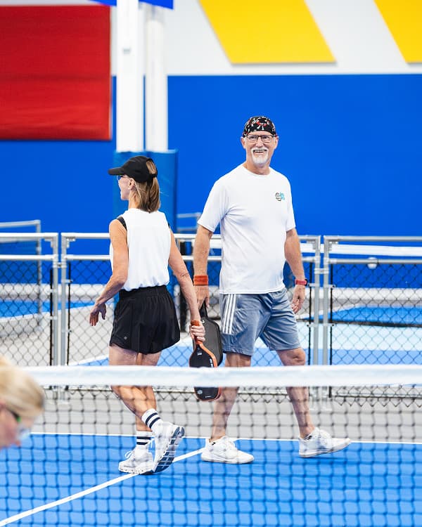 A man and woman walking together on an indoor pickleball court, both smiling and holding paddles.