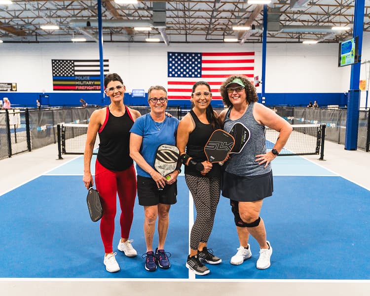 Four women posing on a pickleball court with paddles, in front of large American flags on the wall.