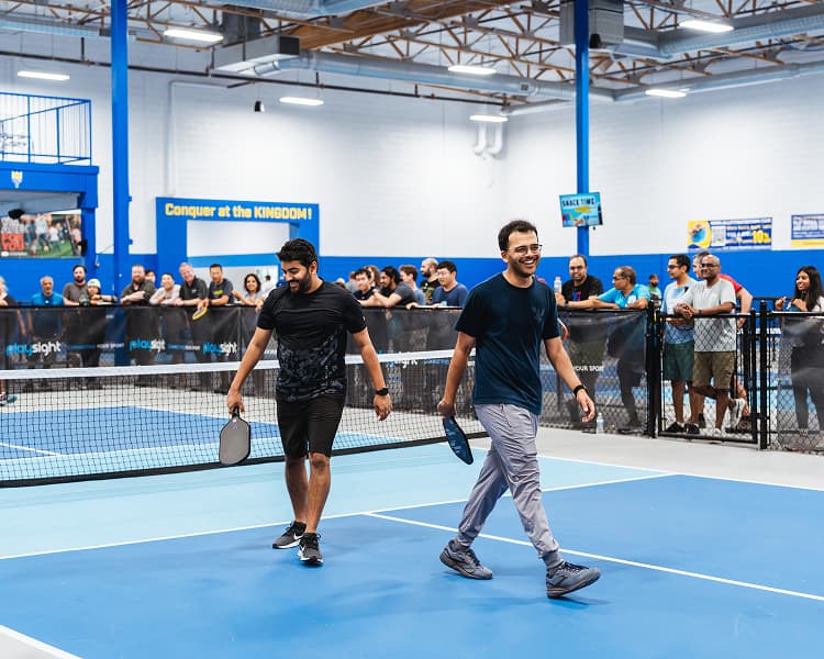 Two men walking off the court smiling after a match, with a crowd watching from behind the fence.