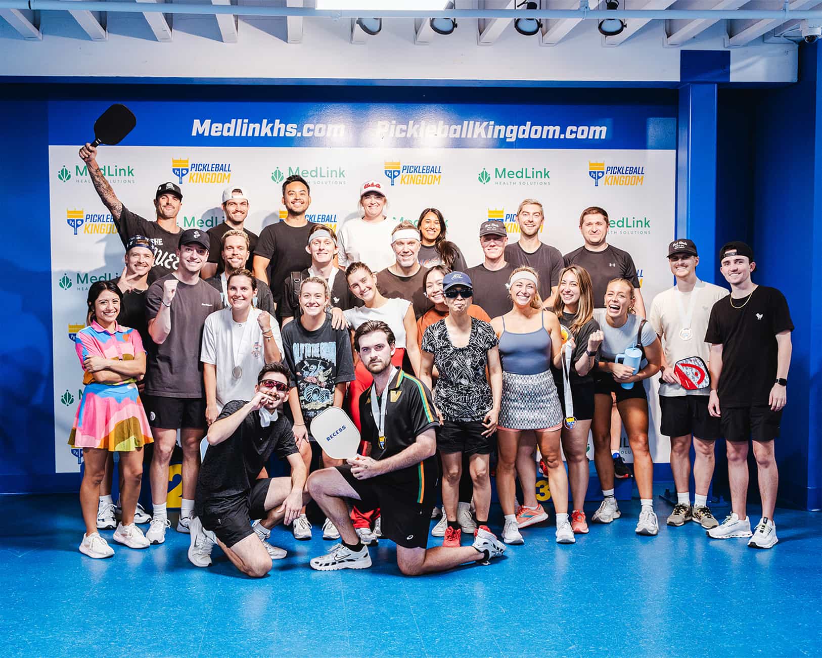 A large group of adults smiling and posing in front of a Pickleball Kingdom sponsor backdrop.